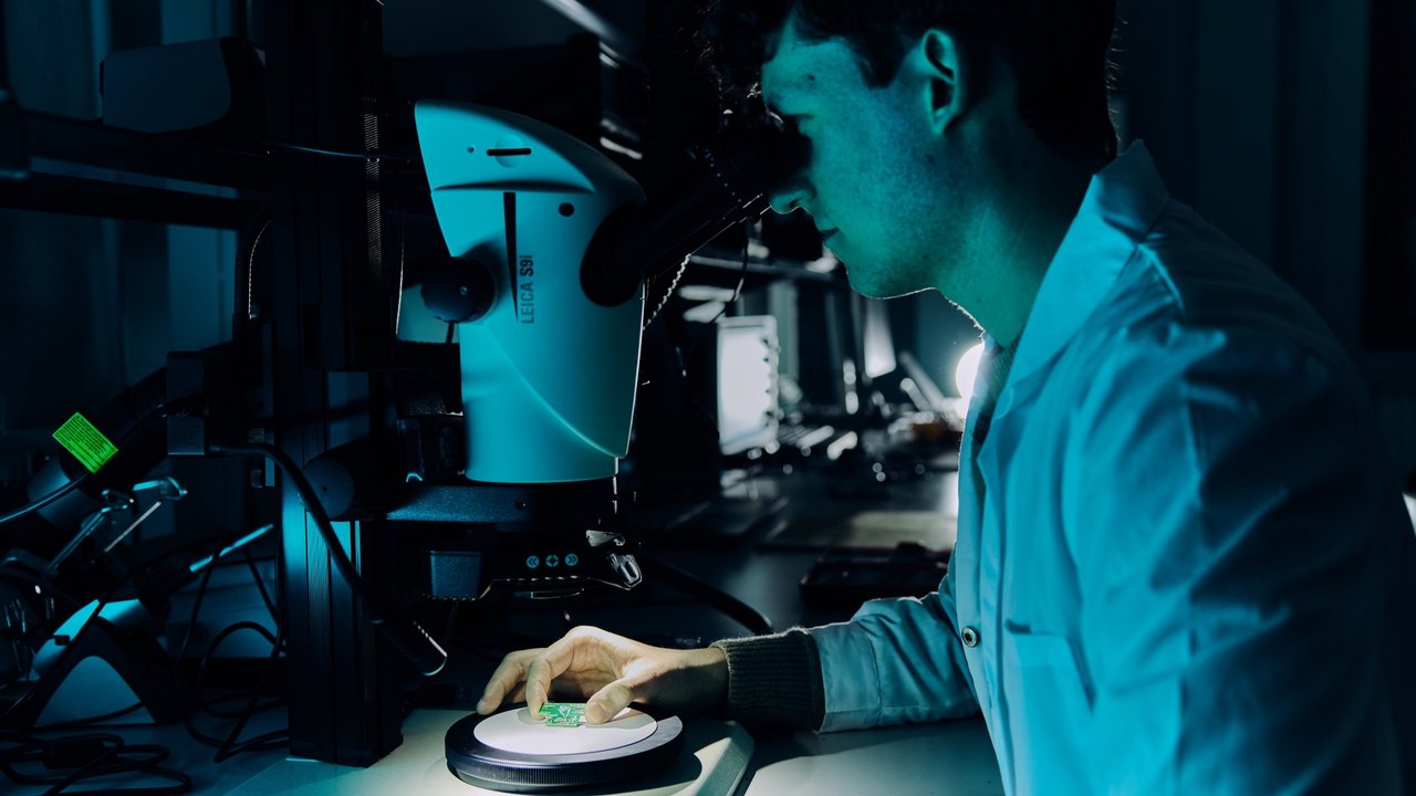 Un ricercatore osserva componenti elettronici e schede attraverso un microscopio, esaminando i dettagli costruttivi e le connessioni durante un’analisi di laboratorio.