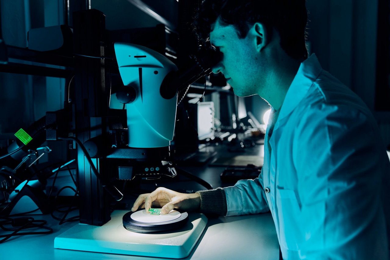 Un ricercatore osserva componenti elettronici e schede attraverso un microscopio, esaminando i dettagli costruttivi e le connessioni durante un’analisi di laboratorio.