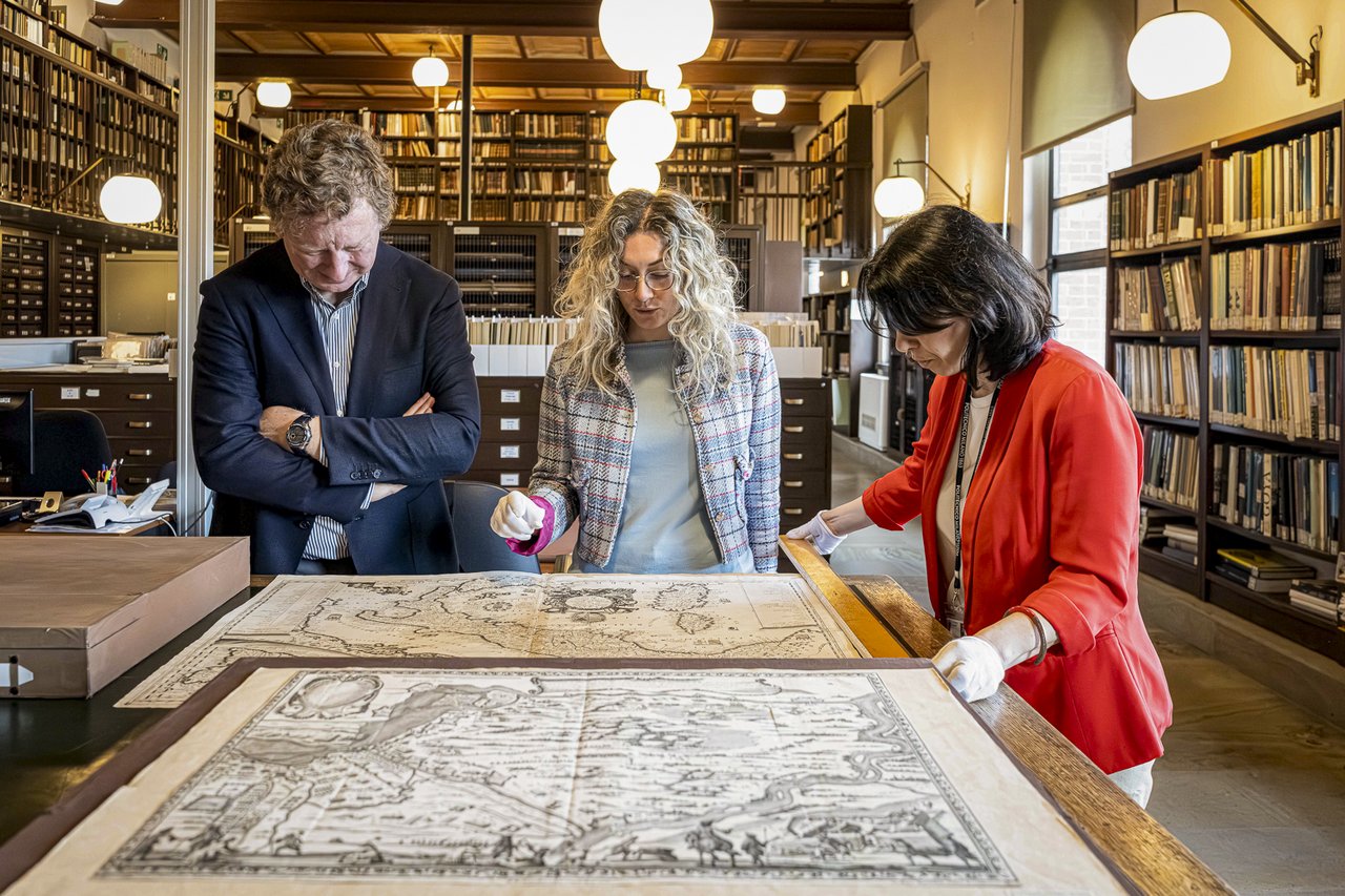 Paolo Bossi, Claudia Giorgi, and Jessica Gritti, from left, examine a historical map spread out on a table in the archive. In the background, shelves and cabinets house volumes and materials from the collection.