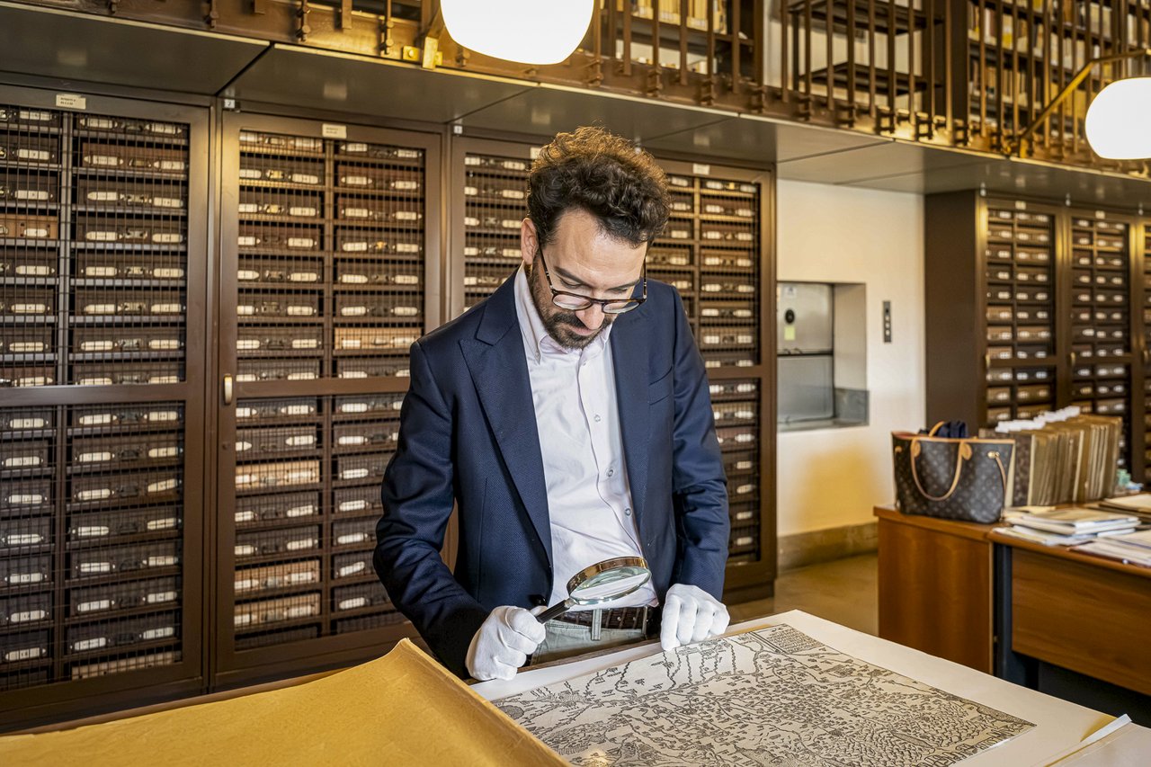Lorenzo Mascheretti examines a historical map spread out on the table with the aid of a magnifying glass, wearing gloves to handle the document. In the background, the archive cabinets house materials from the collection.