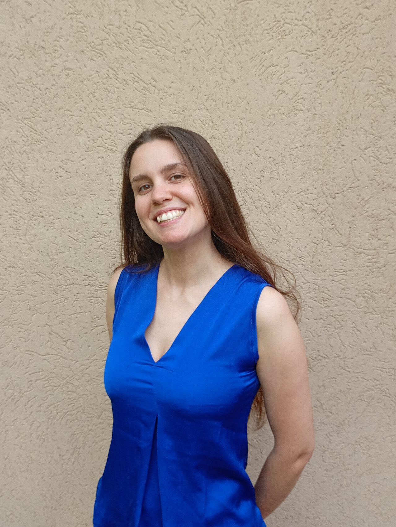 Donna sorridente con lunghi capelli castani, indossa un top blu senza maniche ed è in posa davanti a un muro beige dalla superficie ruvida.