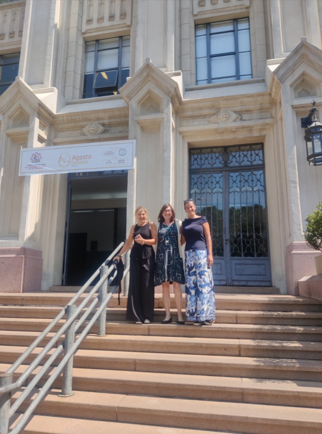 Three people stand smiling on the steps in front of the monumental entrance of the University of São Paulo, Brazil, with large windows, an iron gate, and a banner hanging on the façade.